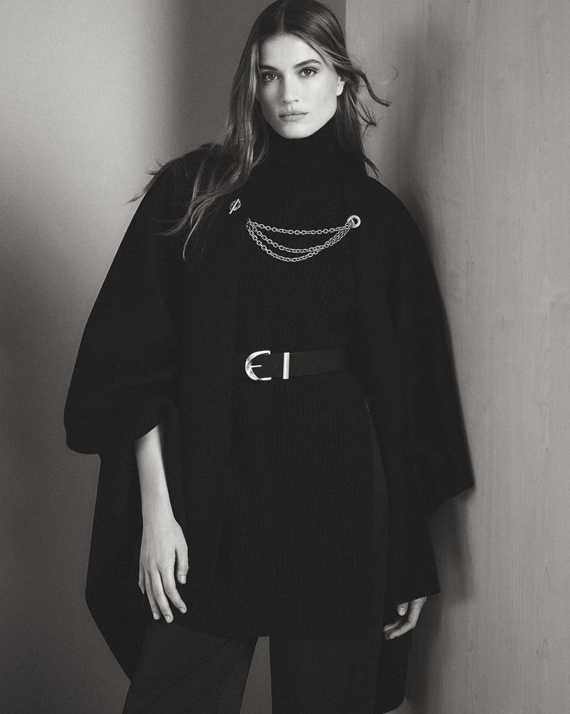 Monochrome portrait of an adult female fashion model in a black turtleneck and black poncho, accessorized with a belt and chains.