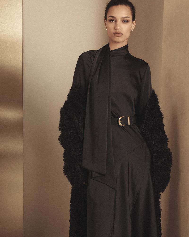A woman in a black dress and faux fur coat poses indoors against a neutral wall with a gold accent, looking directly at the camera.