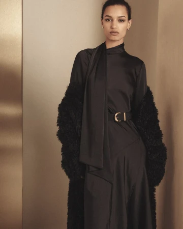 A woman in a black dress and faux fur coat poses indoors against a neutral wall with a gold accent, looking directly at the camera.
