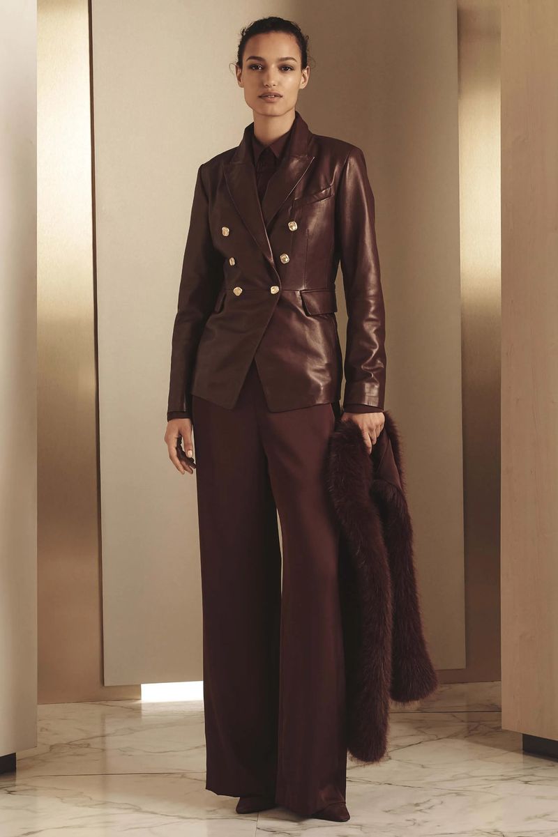 Woman in a burgundy leather double-breasted blazer and matching wide-leg trousers, holding a faux fur stole. Studio lighting.