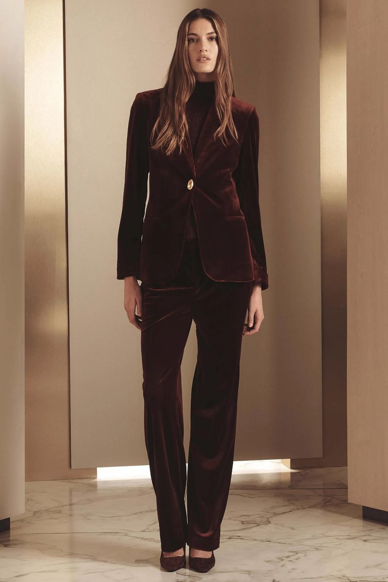 A full-length shot of an adult female model in a burgundy velvet blazer and pants outfit posing indoors in a studio with neutral tones.