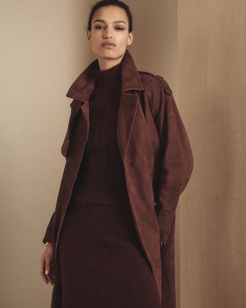 A woman poses in a burgundy sweater dress and suede trench coat indoors, showcasing fashion and style in a studio-lit setting.