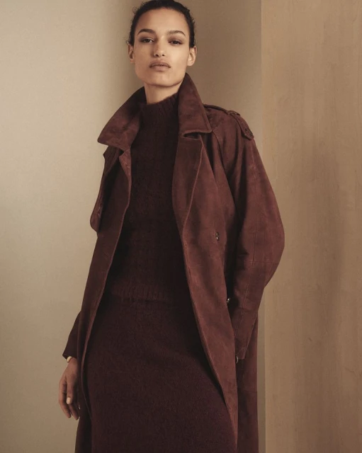 A woman poses in a burgundy sweater dress and suede trench coat indoors, showcasing fashion and style in a studio-lit setting.