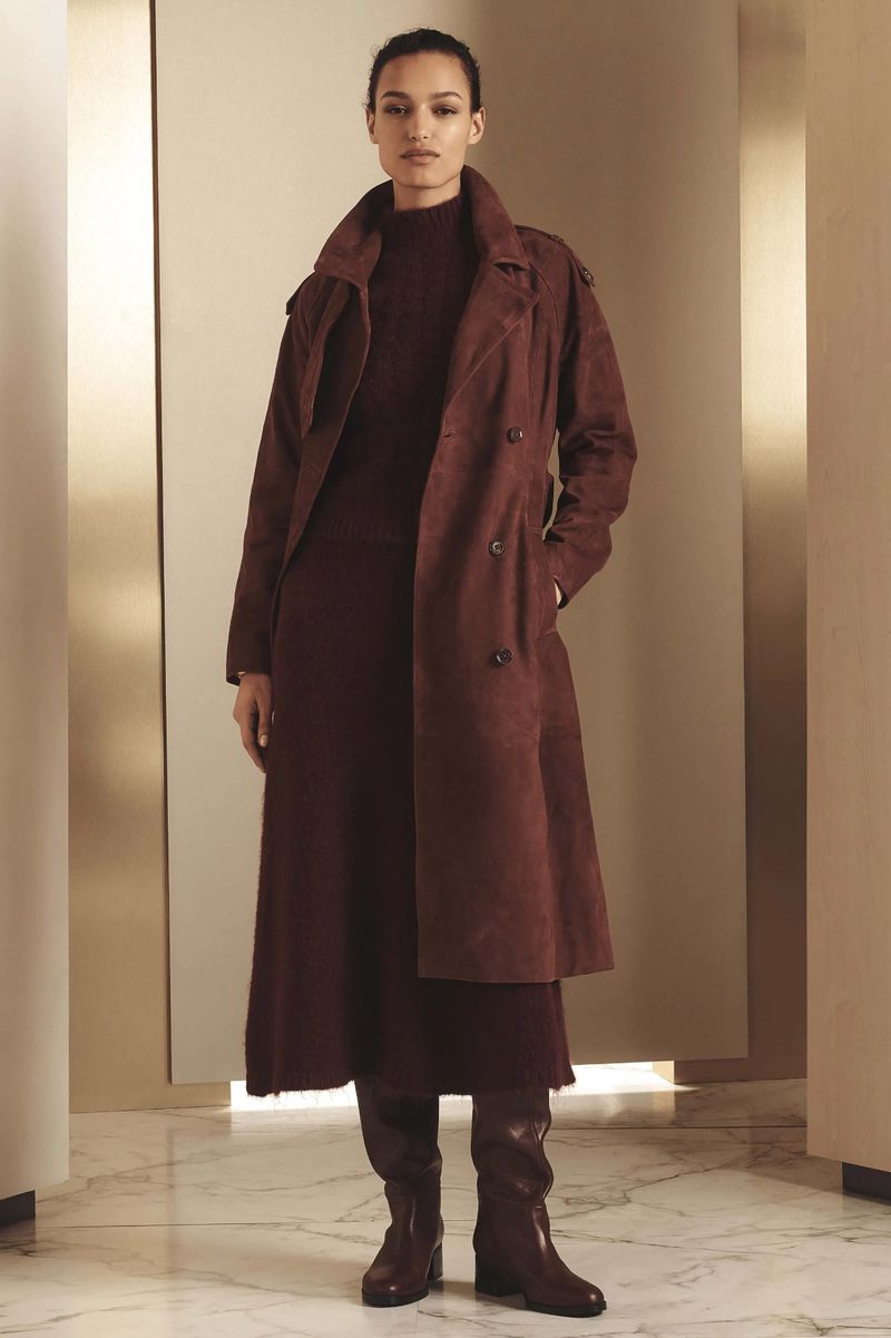 A tall woman poses indoors wearing a brown double-breasted trench coat, dress, and matching knee-high boots against a neutral backdrop.