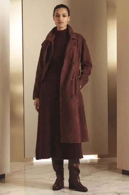 A tall woman poses indoors wearing a brown double-breasted trench coat, dress, and matching knee-high boots against a neutral backdrop.