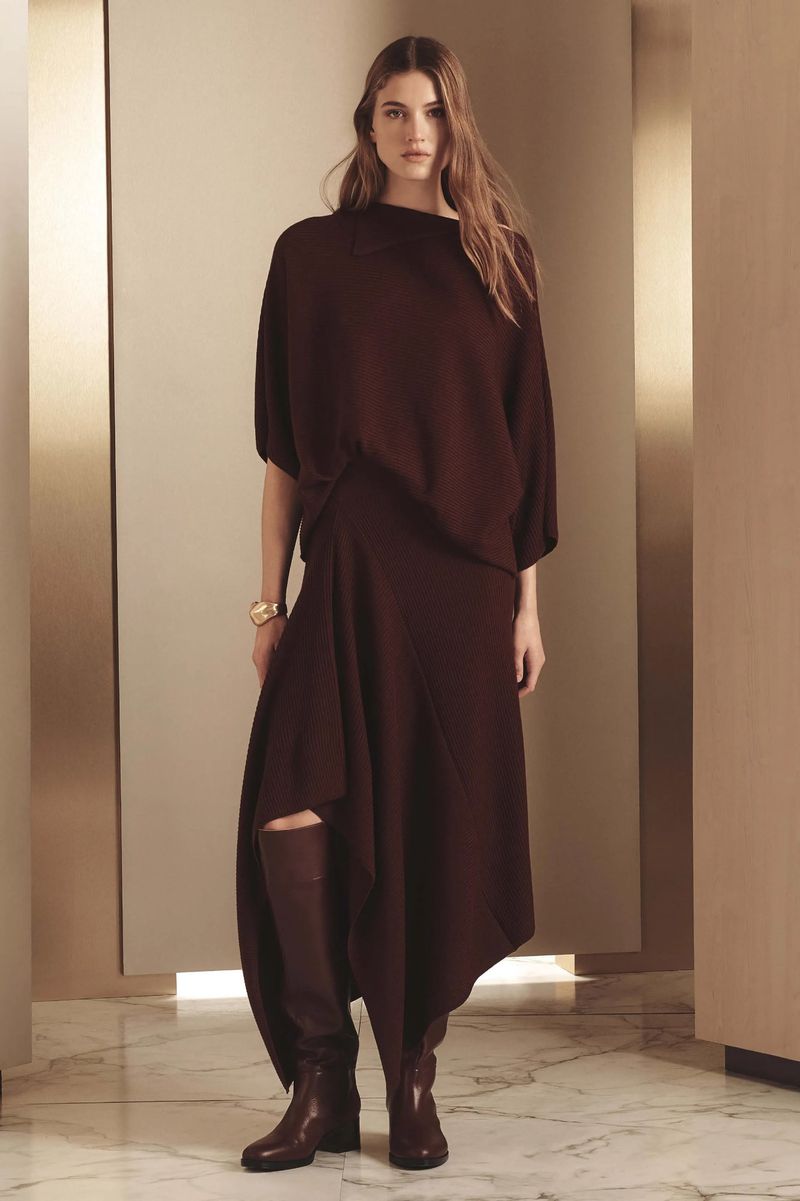 A woman poses indoors wearing a long-sleeved burgundy top, a skirt with an asymmetrical hem, and burgundy knee-high boots for a fashion shot.