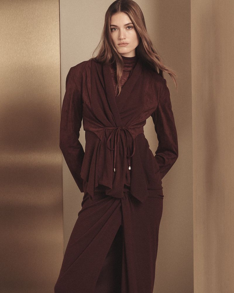 A woman with long brown hair poses indoors, wearing a long-sleeve maroon wrap top and a matching long skirt with a split.