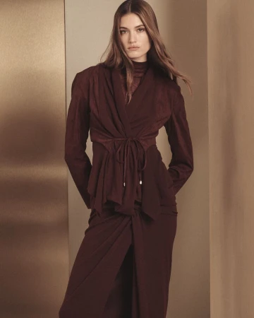 A woman with long brown hair poses indoors, wearing a long-sleeve maroon wrap top and a matching long skirt with a split.