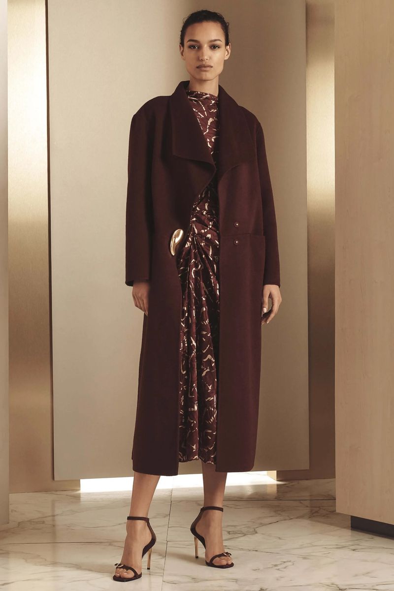 Fashion model in a maroon coat and patterned dress, posing in a studio with a marble floor and tan walls. Elegant and stylish.