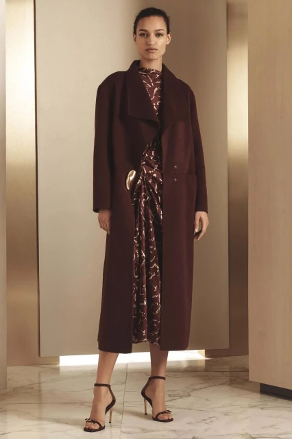Fashion model in a maroon coat and patterned dress, posing in a studio with a marble floor and tan walls. Elegant and stylish.