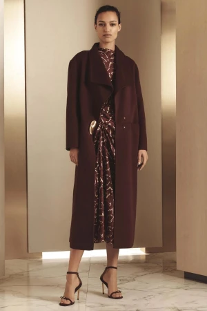Fashion model in a maroon coat and patterned dress, posing in a studio with a marble floor and tan walls. Elegant and stylish.
