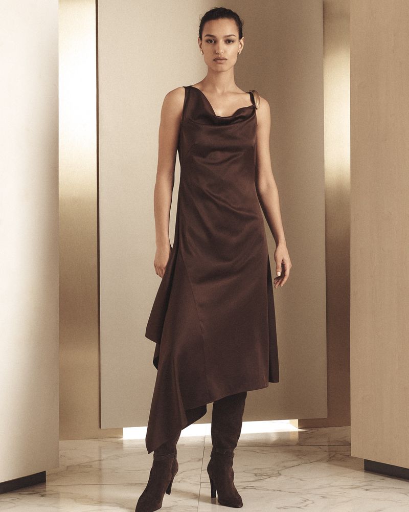 Fashion shoot of a woman in a brown asymmetrical satin dress and knee-high boots in a neutral-toned studio setting with soft lighting.
