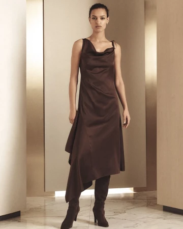 Fashion shoot of a woman in a brown asymmetrical satin dress and knee-high boots in a neutral-toned studio setting with soft lighting.