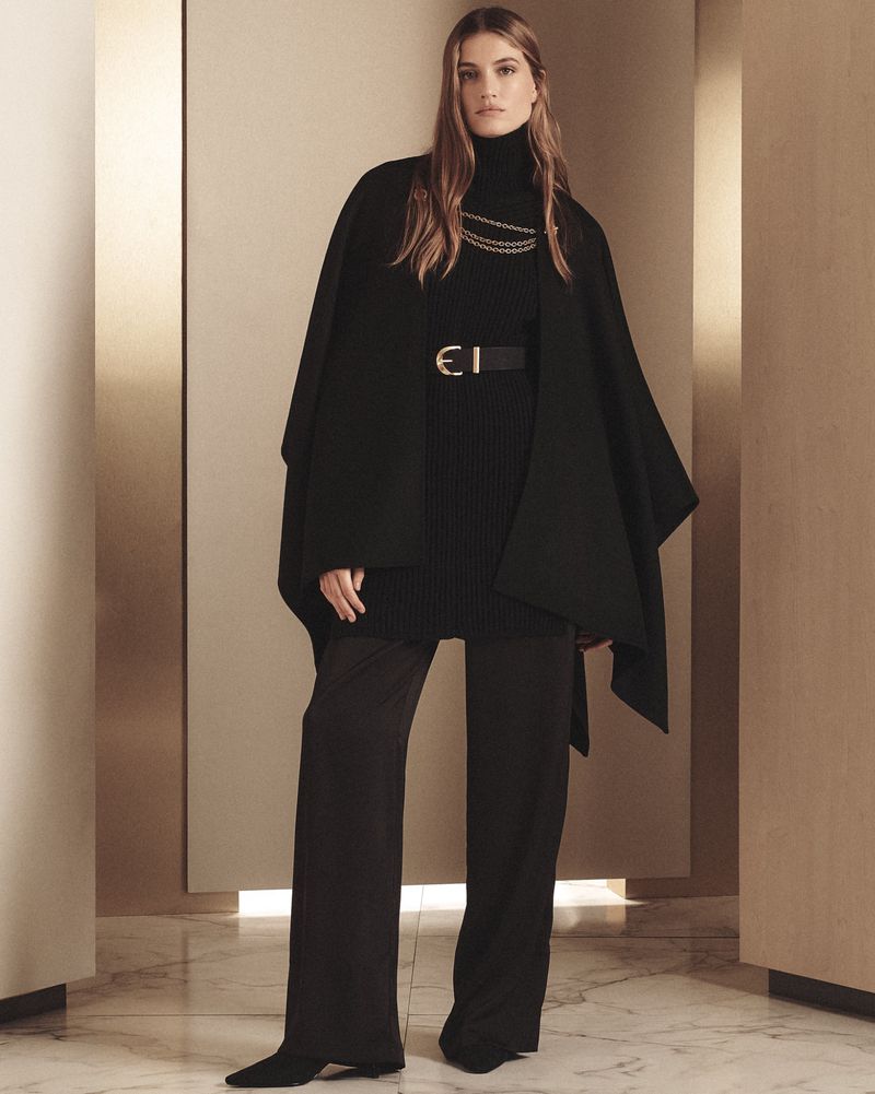 Elegant woman in black turtleneck, cape, wide-leg pants, gold chain, and belt stands in a neutral-toned interior for a fashion editorial.