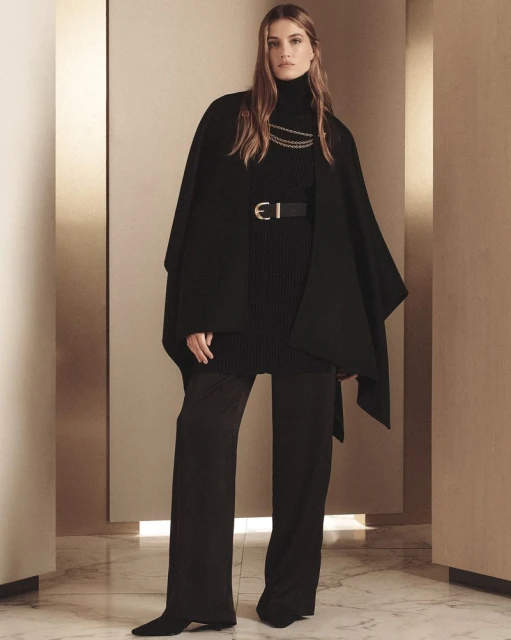 Elegant woman in black turtleneck, cape, wide-leg pants, gold chain, and belt stands in a neutral-toned interior for a fashion editorial.
