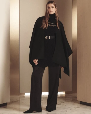 Elegant woman in black turtleneck, cape, wide-leg pants, gold chain, and belt stands in a neutral-toned interior for a fashion editorial.