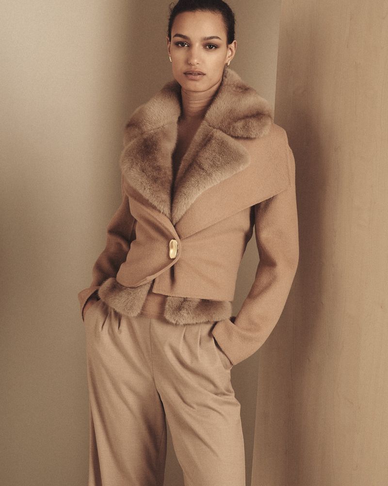 Model wearing a tan fur collar jacket and matching trousers standing against a warm neutral studio wall