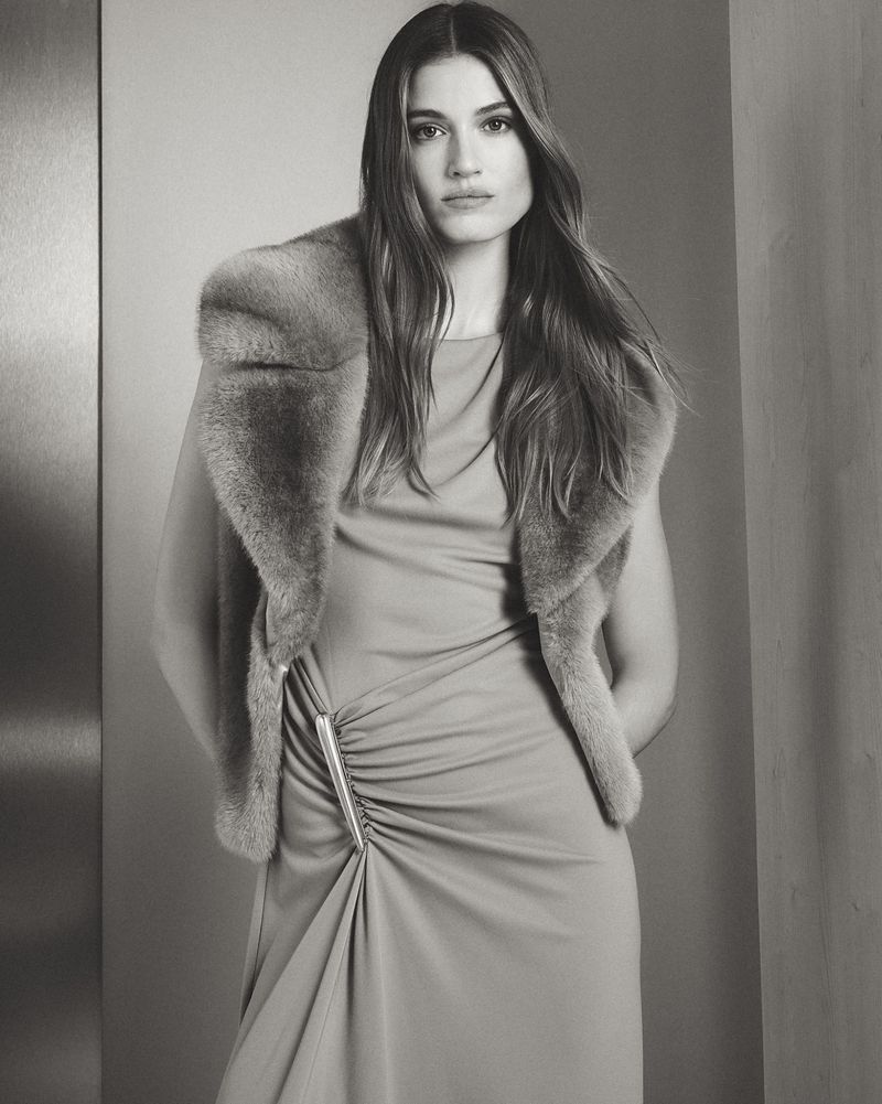 Portrait of a woman wearing a dress and fur stole, posing indoors in a neutral-toned setting with natural light. Fashion advertisement.