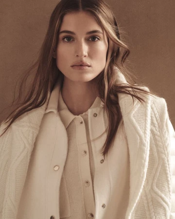 Model wearing a white knitted sweater and draped coat against a textured brown studio background.