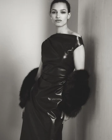 Model in a black glossy dress with fur cuffs leaning against a plain wall in a monochrome portrait.