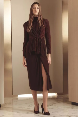 Model wearing a dark burgundy tied jacket and midi skirt with a thigh slit against a minimalist architectural background.