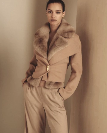 Model wearing a tan fur collar jacket and matching trousers standing against a warm neutral studio wall