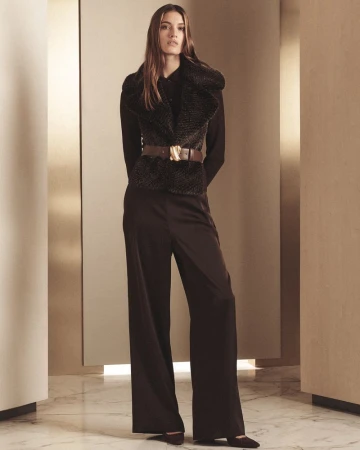 Model standing in dark trousers and a textured vest with a thick brown belt against a neutral architectural backdrop.