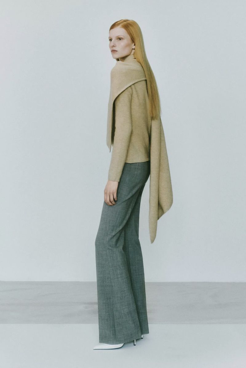 A poised fashion model showcases a tan sweater with draping and gray wide-leg trousers against a plain studio backdrop, radiating elegance.
