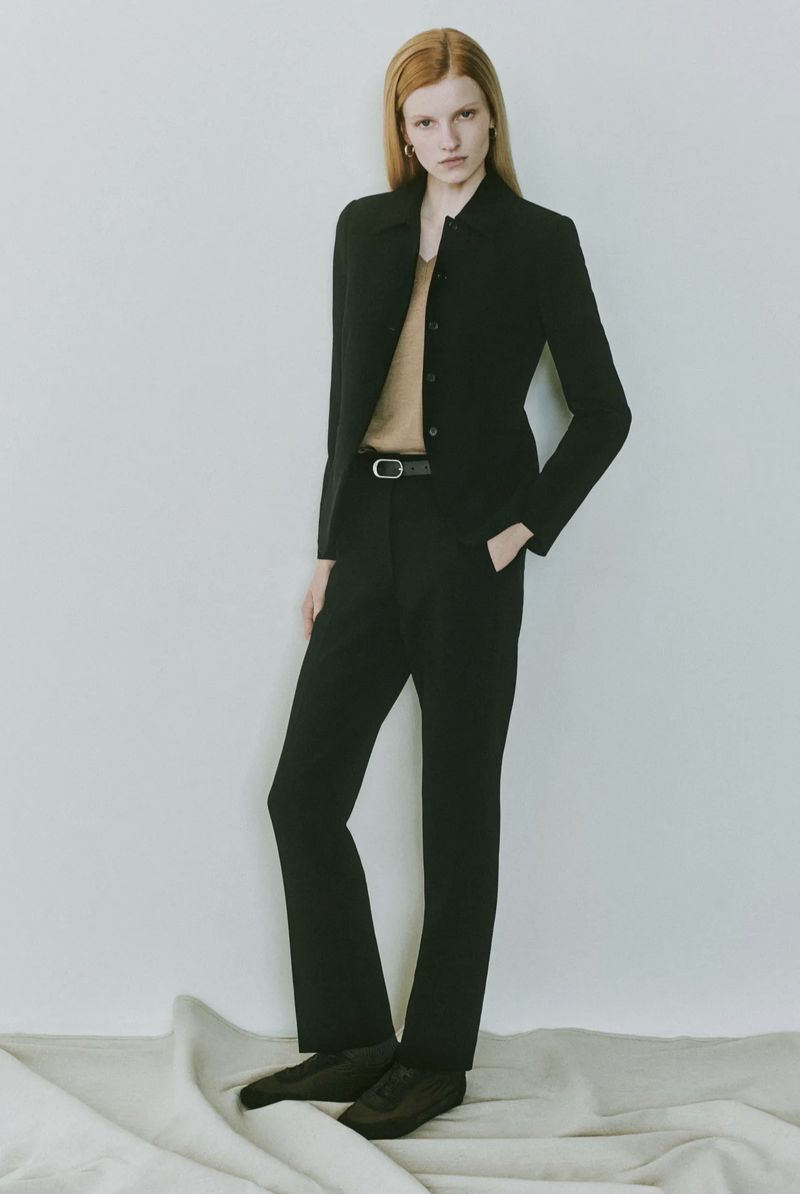 Full-length shot of a red-haired woman in a black blazer and pants, standing on textured white fabric against a white background.