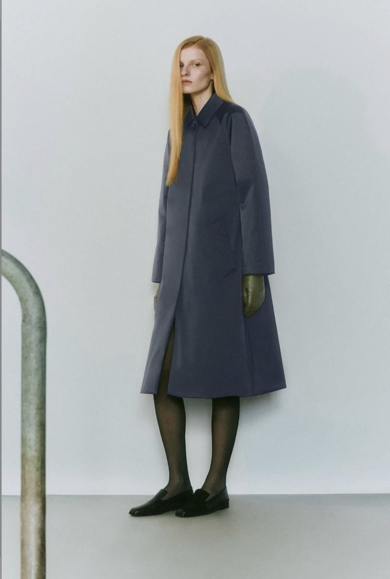 Adult woman stands in front of a gray wall wearing a dark coat, black tights, and black loafers with a curved metal bar visible.