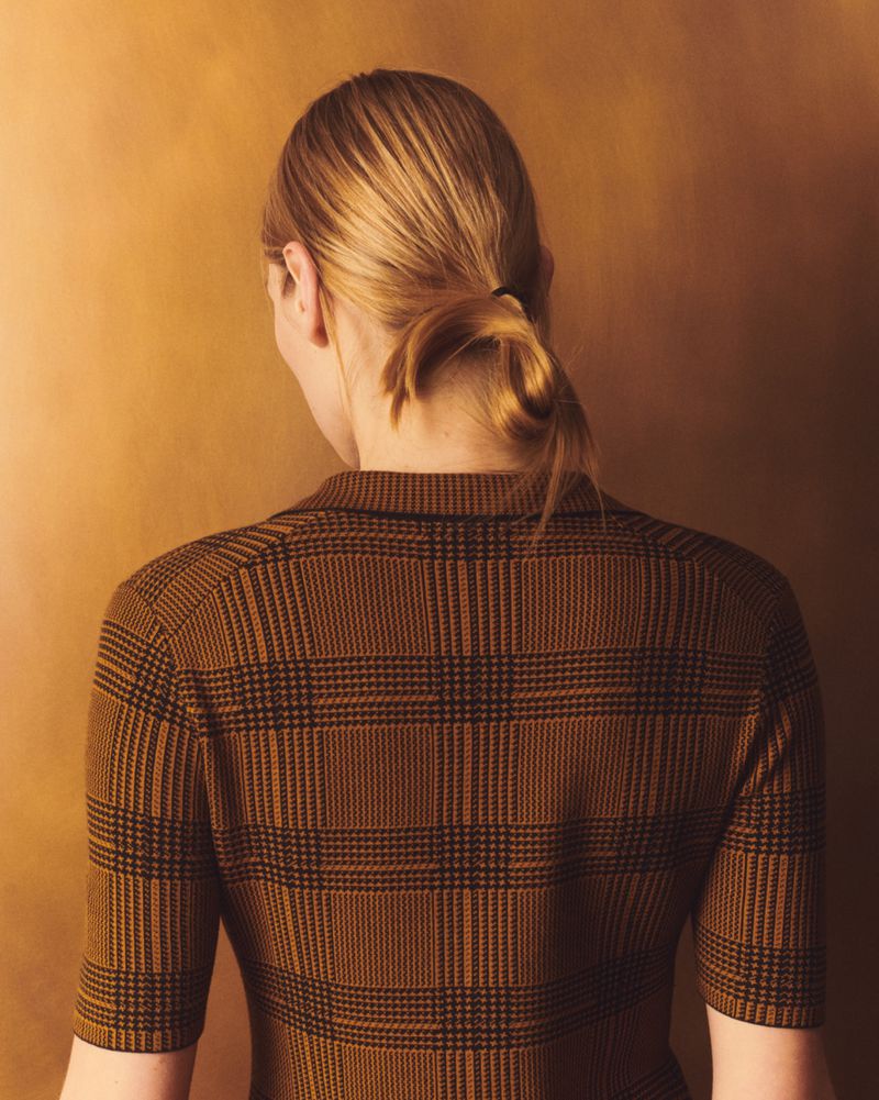 Back view of a blond woman in a brown plaid shirt. The model has a low ponytail and is standing in front of a golden-brown backdrop.