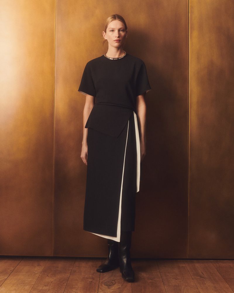 Elegant woman in a black top and wrap skirt stands against a textured gold background in a studio shot, showcasing a modern fashion style.