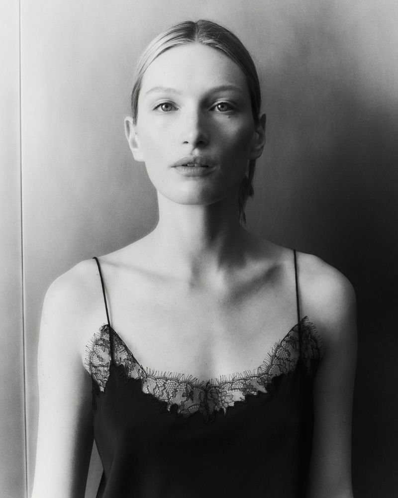 Black and white portrait of a blonde-haired woman in a lace-trimmed camisole against a blurred neutral background, medium shot.