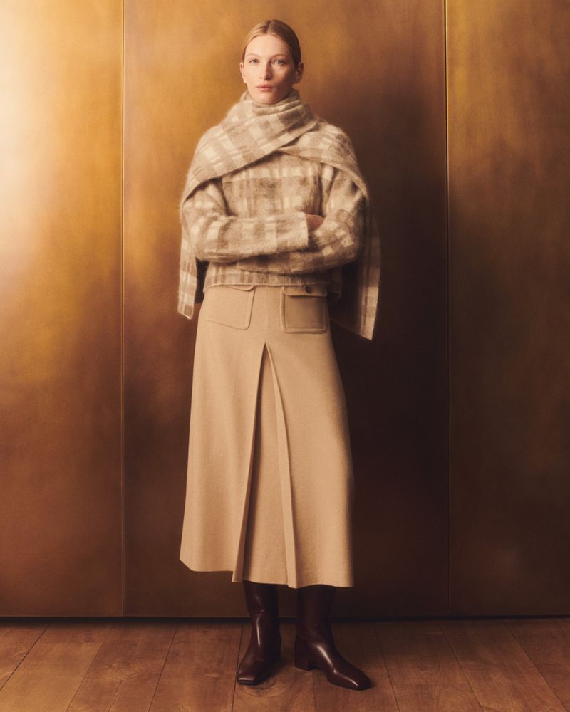 Woman poses in a plaid sweater, scarf, pleated skirt, and brown boots against a golden background for a fashion editorial.