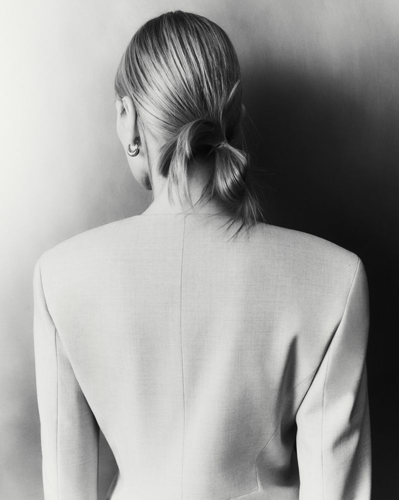Back view of a blonde woman with a low ponytail wearing a minimalist blazer against a textured grey background.