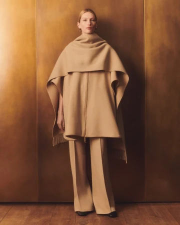 Model wearing a camel wool cape and trousers standing against a warm textured bronze wall for Lafayette 148