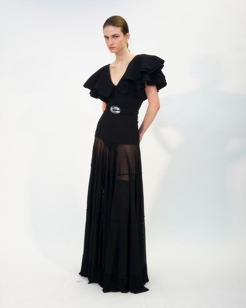 Model wearing a black ruffled evening gown with sheer lower skirt and belt against a plain white background