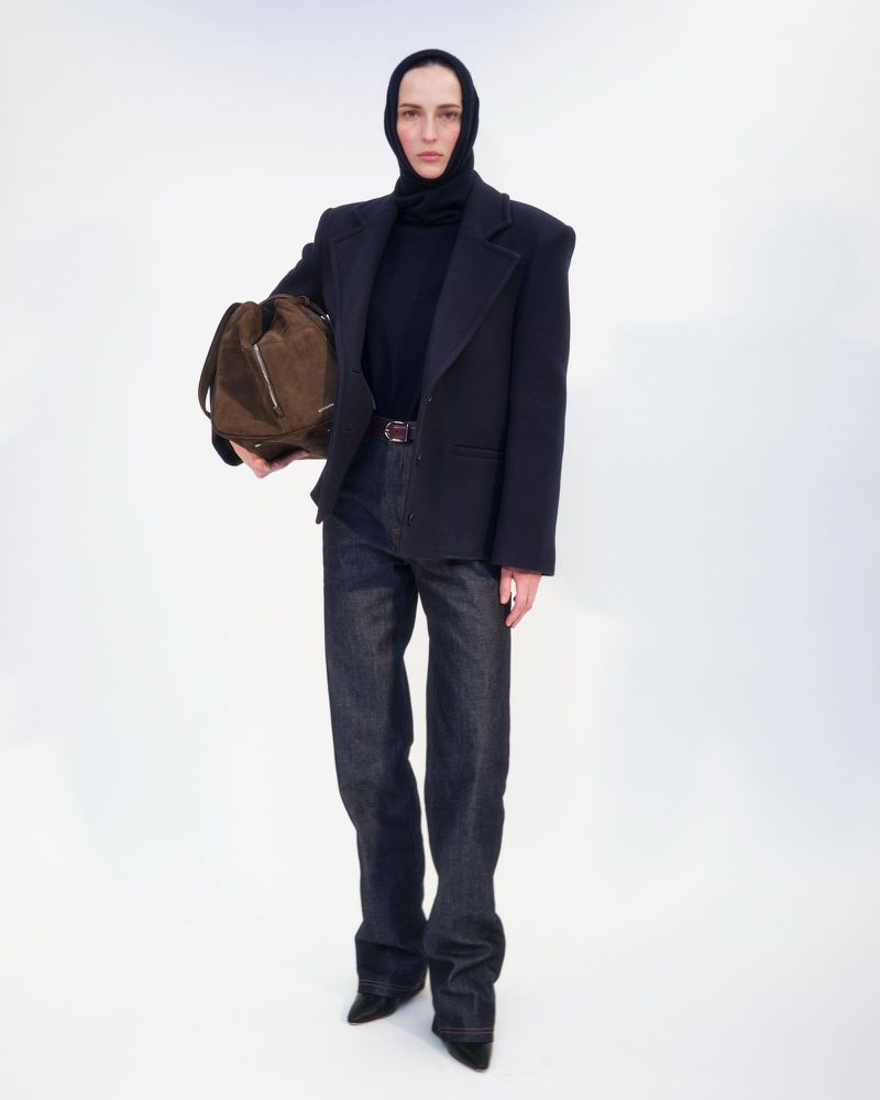 Full-length shot of a young adult female fashion model in a black turtleneck and dark denim, holding a brown bag, on a white backdrop.
