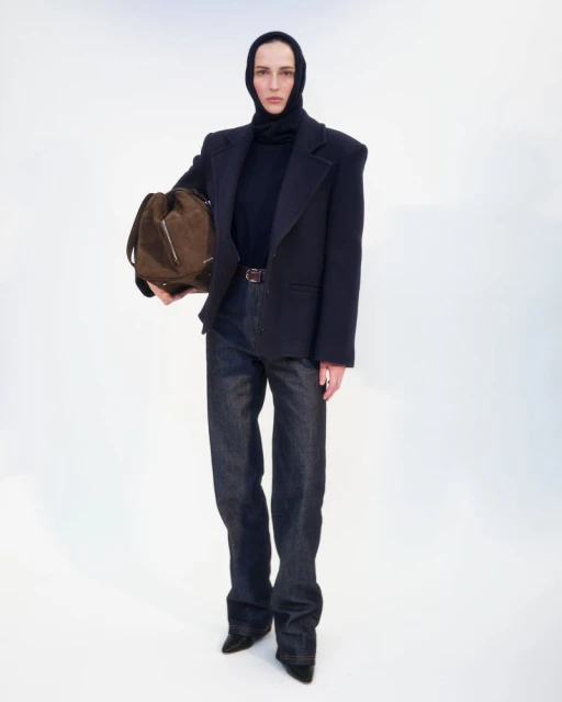 Full-length shot of a young adult female fashion model in a black turtleneck and dark denim, holding a brown bag, on a white backdrop.