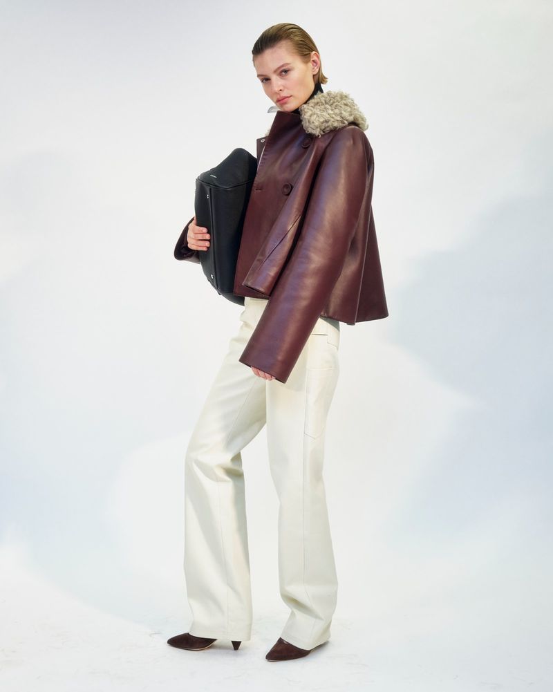 Fashion shot of woman in brown leather jacket and white pants holding black bag, studio lit on white backdrop, stylish pose.