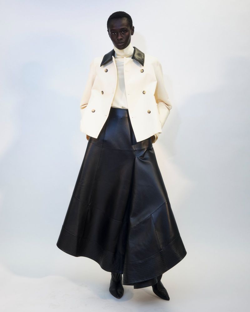 Full-length fashion shot of a Black woman wearing a white jacket, black leather skirt and ankle boots against a neutral background.