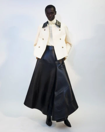 Full-length fashion shot of a Black woman wearing a white jacket, black leather skirt and ankle boots against a neutral background.
