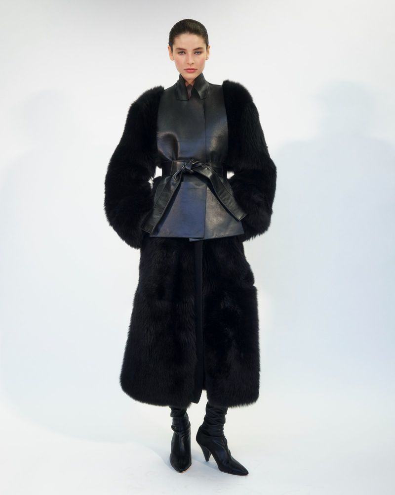 Fashion photo of a woman wearing a black leather top with a belt, long fur coat, pants, and boots against a white studio backdrop.