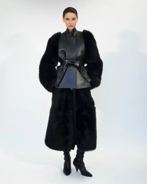 Fashion photo of a woman wearing a black leather top with a belt, long fur coat, pants, and boots against a white studio backdrop.