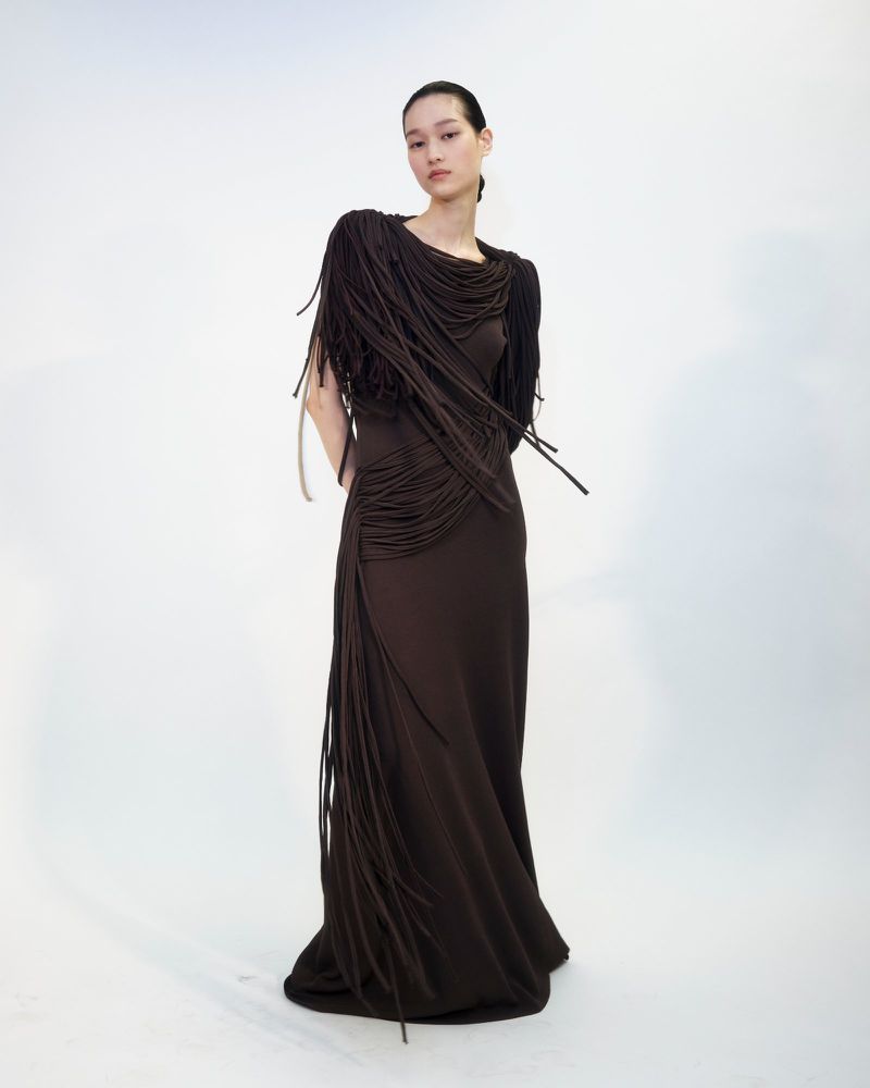 A model stands wearing a long, dark brown dress adorned with cascading fringe details across the shoulders and side against a white background.