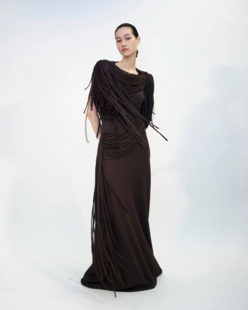 A model stands wearing a long, dark brown dress adorned with cascading fringe details across the shoulders and side against a white background.