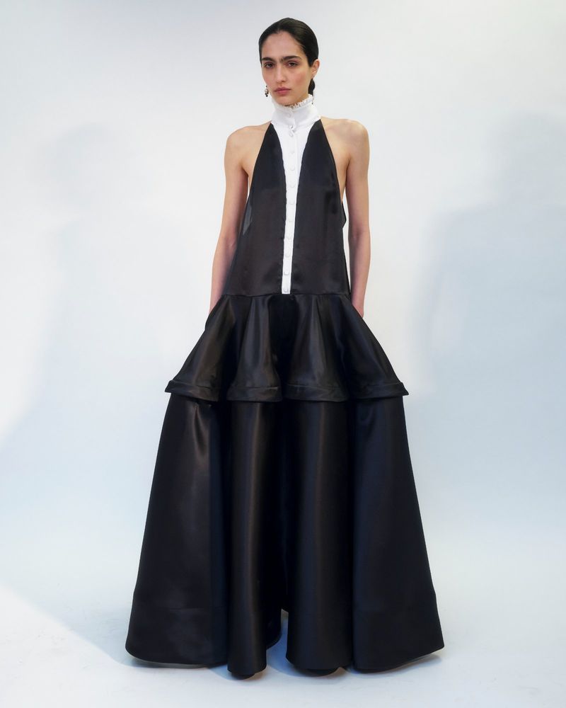 Elegant adult female fashion model in a black sleeveless gown with white halter-style collar posing in a white photo studio.