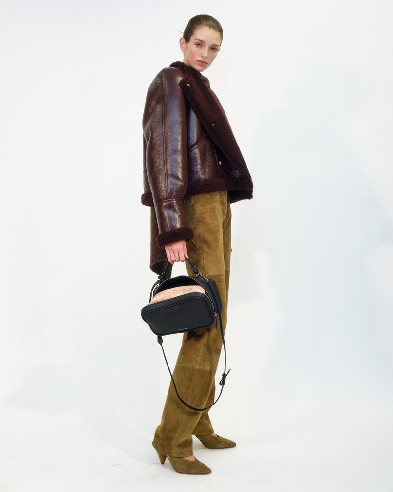 Fashionable woman wearing a brown shearling jacket and olive-green suede pants, carrying a structured black handbag in a studio setting.