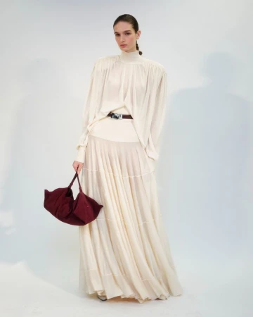 Model in a long cream pleated skirt and high-neck blouse holding a burgundy handbag against a plain white background.
