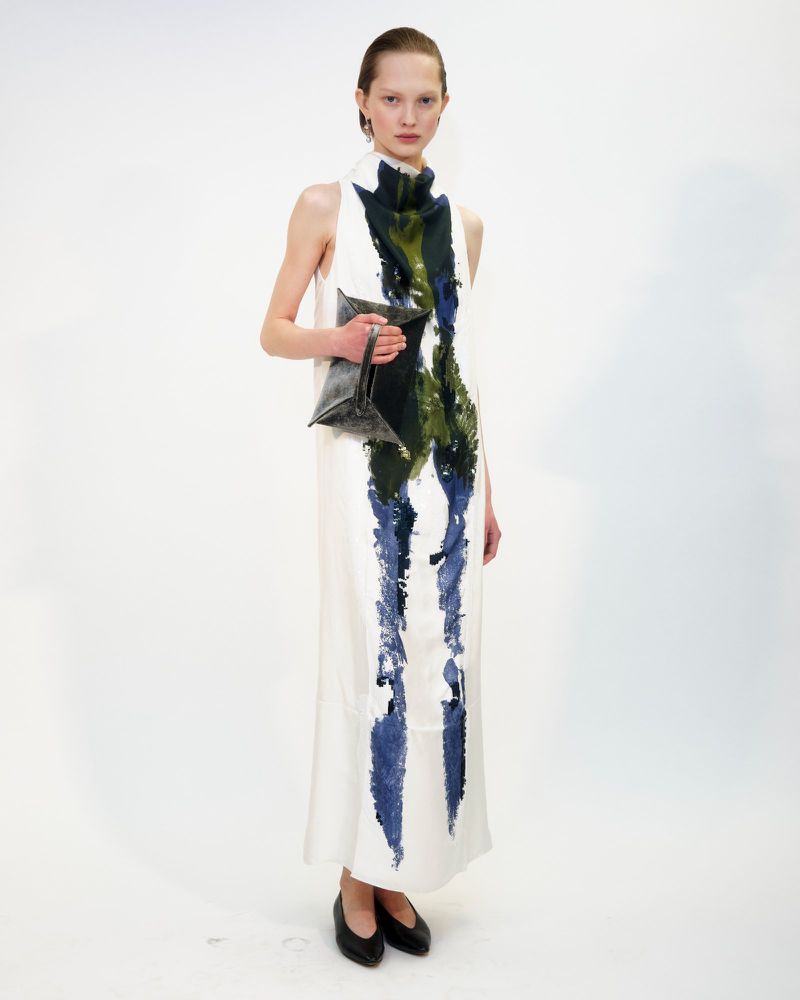 Adult female fashion model poses in a floor-length, sleeveless white dress with green and blue painted accents, holding a geometric handbag.